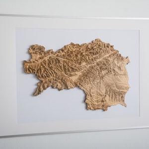 Könnte beinhalten: Eine gerahmte, dreidimensionale Holzreliefkarte einer Bergregion. Die Karte ist in Braun- und Beigetönen gehalten, mit einem weißen Passepartout und einem silbernen Rahmen.