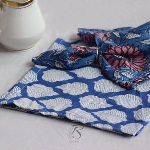 May include: A white ceramic jug with a gold rim is next to two blue cloth napkins. One napkin has a white leaf pattern, and the other has a floral design with pink and blue hues. The napkins are folded.