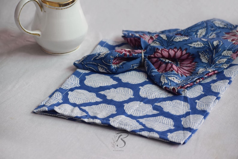 May include: A white ceramic jug with a gold rim is next to two blue cloth napkins. One napkin has a white leaf pattern, and the other has a floral design with pink and blue hues. The napkins are folded.
