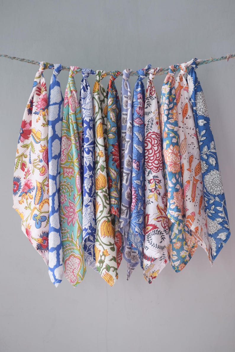 May include: Ten patterned fabric squares in various colours, including blue, pink, and yellow, hang from a rope. The fabrics display floral and geometric designs. The textiles are knotted at the top, creating a colourful and decorative display.