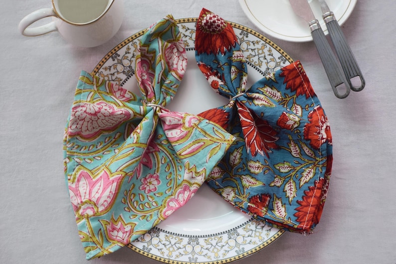 May include: Two floral print cloth napkins, one in teal with pink and yellow flowers, the other in teal with red flowers, are tied with napkin rings and placed on a white plate with a gold rim. A white creamer and silverware are also visible.