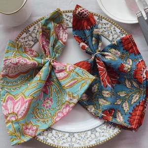 May include: Two fabric napkins with floral patterns. One napkin is blue with red flowers and the other is teal with pink flowers. The napkins are folded and placed on a white plate with a gold rim.