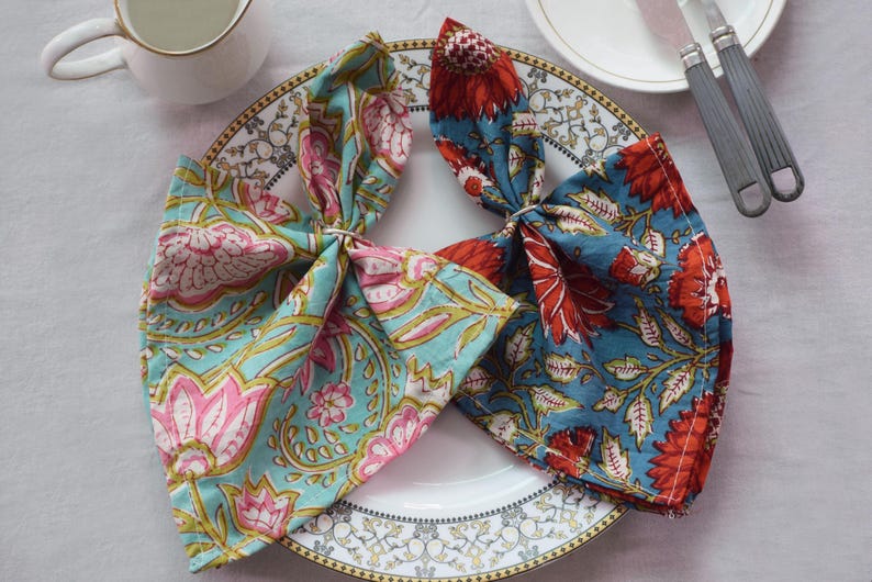 May include: Two fabric napkins with floral patterns. One napkin is blue with red flowers and the other is teal with pink flowers. The napkins are folded and placed on a white plate with a gold rim.
