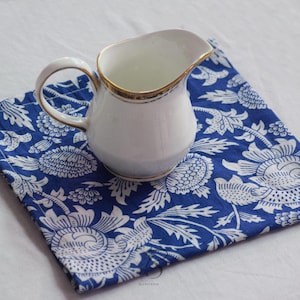 May include: A white ceramic creamer with a gold rim and handle rests on a blue and white floral patterned cloth. The cloth is square and folded. The creamer is empty.