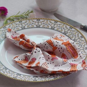 May include: A white dinner plate with a gold and gray patterned border. A white napkin with orange and pink fish designs is folded on the plate.