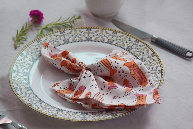 May include: A white dinner plate with a gold and gray patterned border. A white napkin with orange and pink fish designs is folded on the plate.