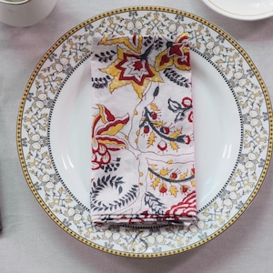 May include: A white plate with a gold and black patterned rim holds a folded napkin with a floral design in red, yellow, and black. A silver fork and knife with grey handles are on either side.