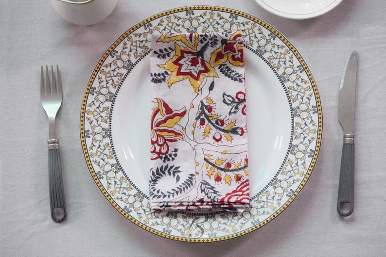 May include: A white plate with a gold and black patterned rim holds a folded napkin with a floral design in red, yellow, and black. A silver fork and knife with grey handles are on either side.