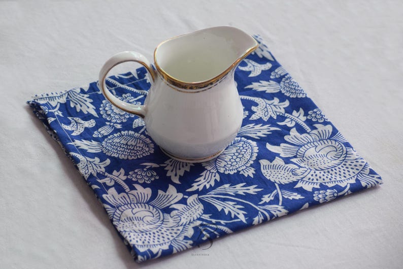 May include: A white ceramic creamer with a gold trim sits on a blue and white floral patterned fabric napkin.
