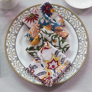 May include: Four folded fabric napkins with floral patterns in shades of blue, yellow, red, and white. The napkins are arranged on a white plate with a gold rim.