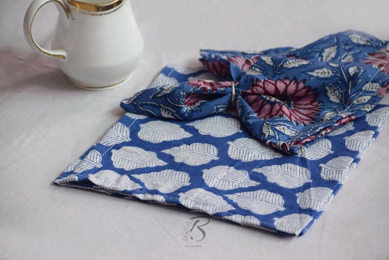 May include: Two blue and white cotton napkins with a floral print. The top napkin is folded into a triangle shape and has a pink and blue floral design. The bottom napkin has a white leaf pattern on a blue background.