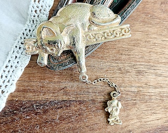 Op de afbeelding: Gouden kattenhanger met een ketting en een kleinere hanger van een kat. De kattenhanger zit op een decoratieve basis.