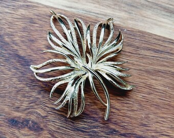 May include: A gold-toned metal brooch in the shape of a stylized leaf. The leaf has a textured surface and is detailed with intricate lines.