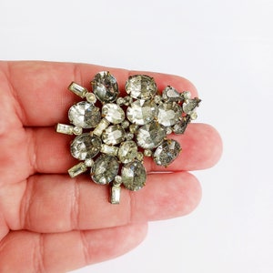 May include: A silver tone brooch with a cluster of clear and gray rhinestones in a flower shape.