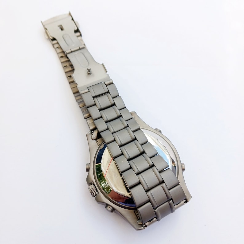 May include: A silver-toned wristwatch with a metal strap. The watch is laid flat on a white surface with the strap folded over the back of the watch.