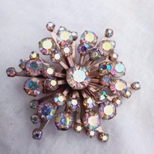 K&ouml;nnte beinhalten: Eine Vintage-Brosche mit einem Strahlenkranz aus schillernden Strasssteinen in Rosa-, Lila- und Gelbt&ouml;nen. Die Strasssteine sind in einer goldfarbenen Metallbasis gefasst.