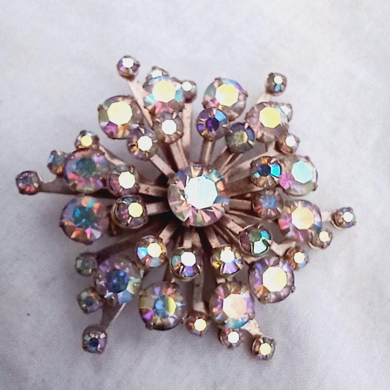 K&ouml;nnte beinhalten: Eine Vintage-Brosche mit einem Strahlenkranz aus schillernden Strasssteinen in Rosa-, Lila- und Gelbt&ouml;nen. Die Strasssteine sind in einer goldfarbenen Metallbasis gefasst.