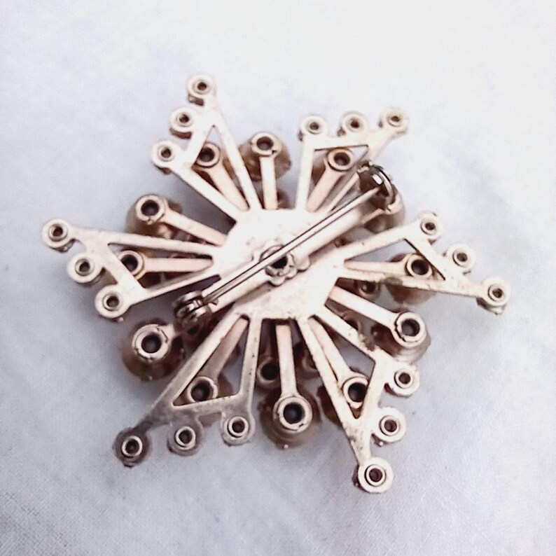 K&ouml;nnte beinhalten: Die R&uuml;ckseite einer silberfarbenen Brosche mit einem Sternburst-Design. Die Brosche ist mit klaren Strasssteinen verziert und hat eine Nadel und einen Sicherheitsverschluss.