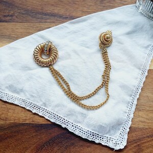 May include: A gold-tone brooch with a circular design and a row of clear rhinestones. The brooch is attached to a double chain.