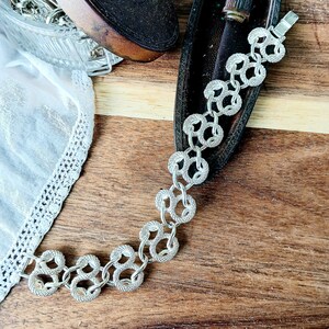 Puede incluir: Un brazalete de cadena de plata con un diseño delicado e intrincado. La cadena está formada por pequeños círculos unidos que crean un aspecto fluido y elegante.