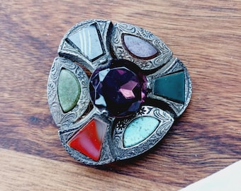 May include: A vintage brooch with a central purple gemstone and surrounding colorful stones in red, green, blue, and gray. The silver-toned metal has an ornate, engraved design. The brooch is circular and set on a wooden surface.