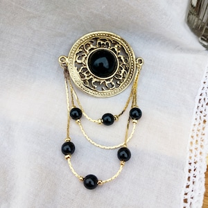 Puede incluir: Un broche dorado con una piedra negra en el centro, rodeado de intrincados detalles de filigrana. El broche está unido a una cadena con cuentas negras.