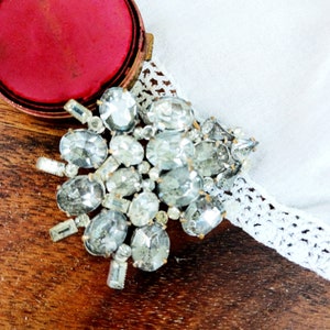 May include: A large silver rhinestone brooch with many faceted stones. The brooch is on a wooden surface with a red velvet box in the background.