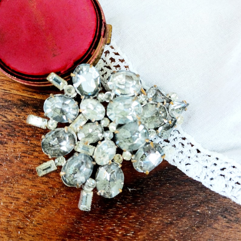 May include: A large silver rhinestone brooch with many faceted stones. The brooch is on a wooden surface with a red velvet box in the background.