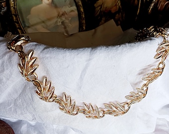May include: A gold-tone necklace with a leaf design. The necklace is on a white fabric background.