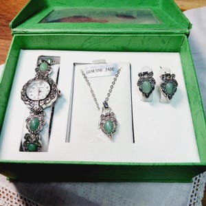 May include: A green gift box containing a matching jewelry set. The set includes a watch, a necklace, and a pair of earrings. The watch and jewelry have silver-colored metalwork and green jade stones. The box is green with a clear window.