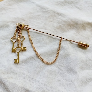 Puede incluir: Un broche dorado con una cadena y tres dijes de llaves. Las llaves son en forma de corazón y tienen un estilo vintage.