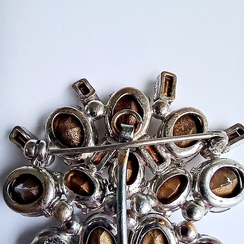 May include: The back of a silver and gold brooch with a cluster of oval-shaped rhinestones. The brooch has a pin and catch closure.