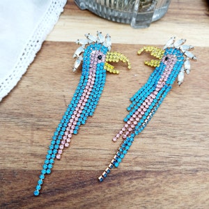 May include: A pair of long, dangling earrings featuring a blue, pink, and yellow bird design with crystal embellishments.