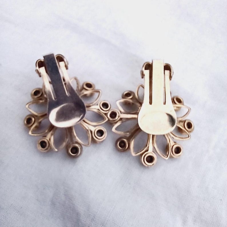 K&ouml;nnte beinhalten: Ein Paar goldfarbene Ohrclips mit einem floralen Design. Die Ohrringe haben einen Cluster aus kleinen, runden Steinen in der Mitte und sind von einem zarten, blattartigen Muster umgeben.