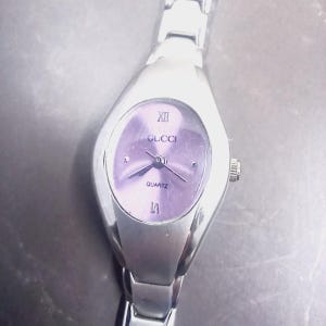 May include: A silver Gucci quartz watch with a lavender oval face. The watch features Roman numeral hour markers and the Gucci logo. The watch band is silver-toned.
