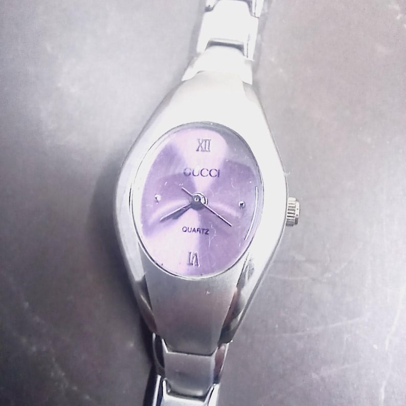 May include: A silver Gucci quartz watch with a lavender oval face. The watch features Roman numeral hour markers and the Gucci logo. The watch band is silver-toned.