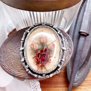May include: A silver-toned oval brooch with a glass dome containing pressed dried flowers. The flowers are pink, white, and red.