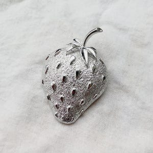 May include: A silver-toned strawberry brooch with a textured surface and a detailed stem and leaves.
