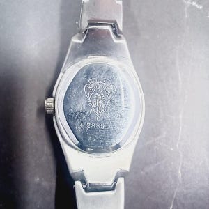 May include: Silver wristwatch with a rectangular face and a matching strap. The watch back displays a logo and the number 28891. The watch has a small, round crown and a polished finish, indicating a classic design.