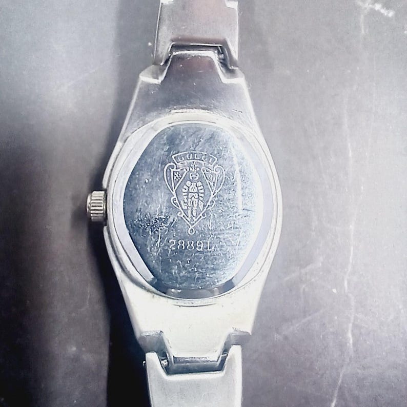 May include: Silver wristwatch with a rectangular face and a matching strap. The watch back displays a logo and the number 28891. The watch has a small, round crown and a polished finish, indicating a classic design.