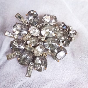 May include: A large, silver-toned brooch with a cluster of clear, faceted rhinestones. The rhinestones are arranged in a random pattern and are of varying sizes.