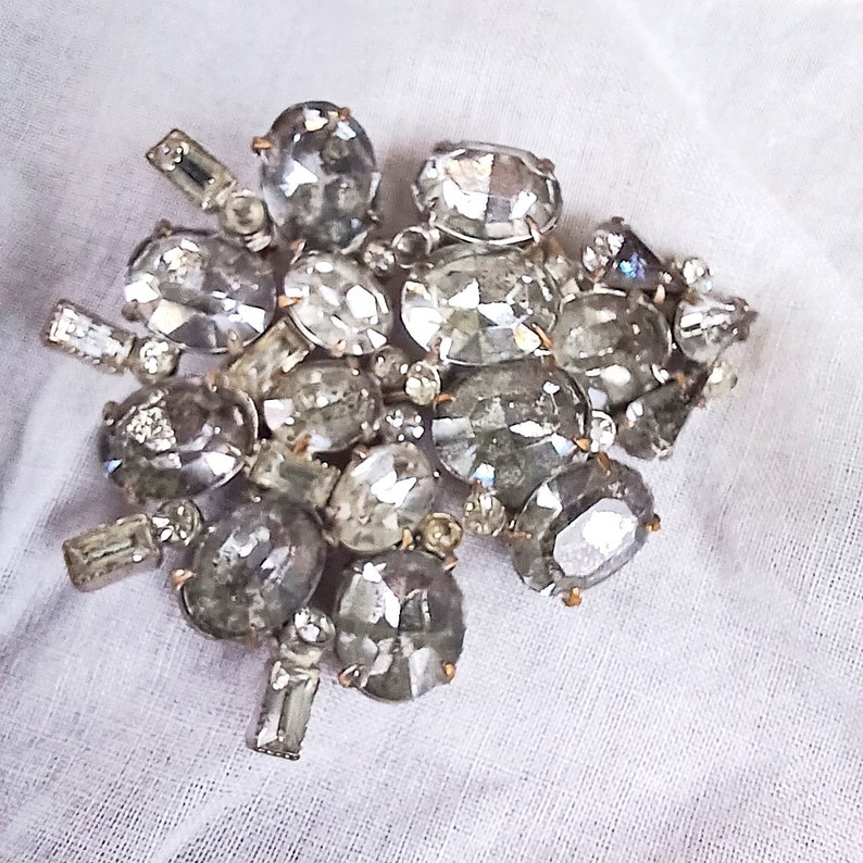 May include: A large, silver-toned brooch with a cluster of clear, faceted rhinestones. The rhinestones are arranged in a random pattern and are of varying sizes.