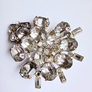 May include: A cluster of clear and silver-toned rhinestones, some oval-shaped and some rectangular, arranged in a random pattern. The rhinestones are attached to a metal base.