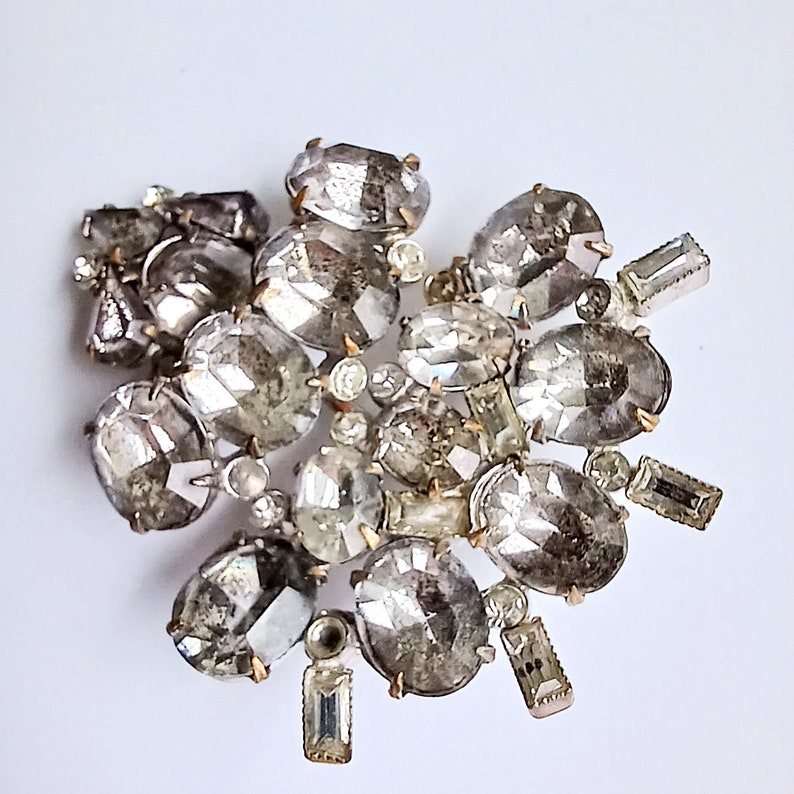 May include: A cluster of clear and silver-toned rhinestones, some oval-shaped and some rectangular, arranged in a random pattern. The rhinestones are attached to a metal base.