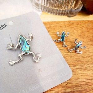 May include: Two silver frog brooches with iridescent blue and green abalone shell inlays. The larger brooch is on a gray card with the text "Island Jewelry".