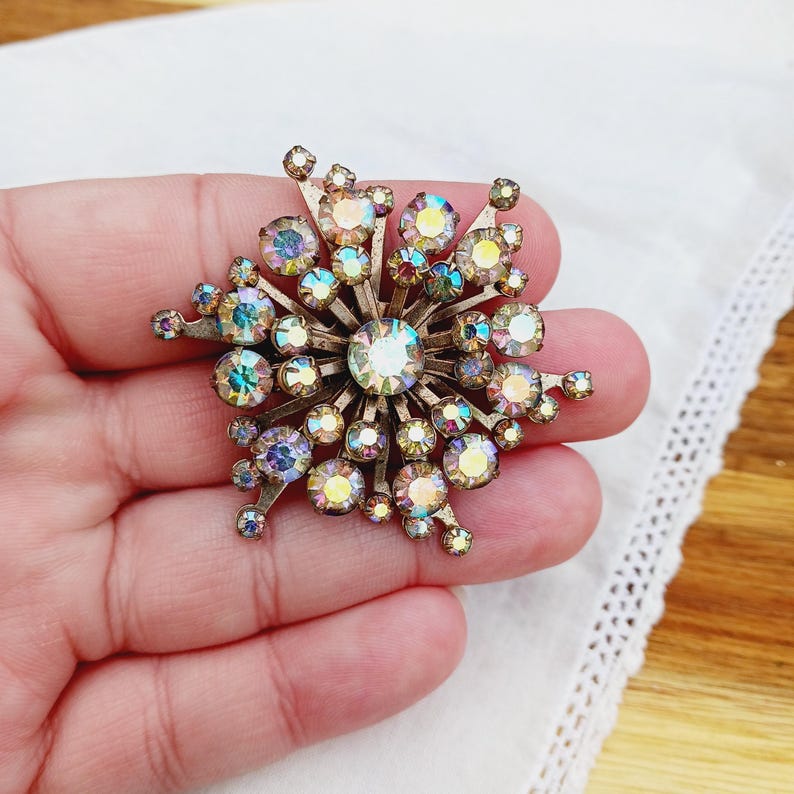 K&ouml;nnte beinhalten: Eine gro&szlig;e, irisierende, goldfarbene Brosche mit einem Sternburst-Design. Die Brosche ist aus Metall und mit kleinen, funkelnden Strasssteinen besetzt.