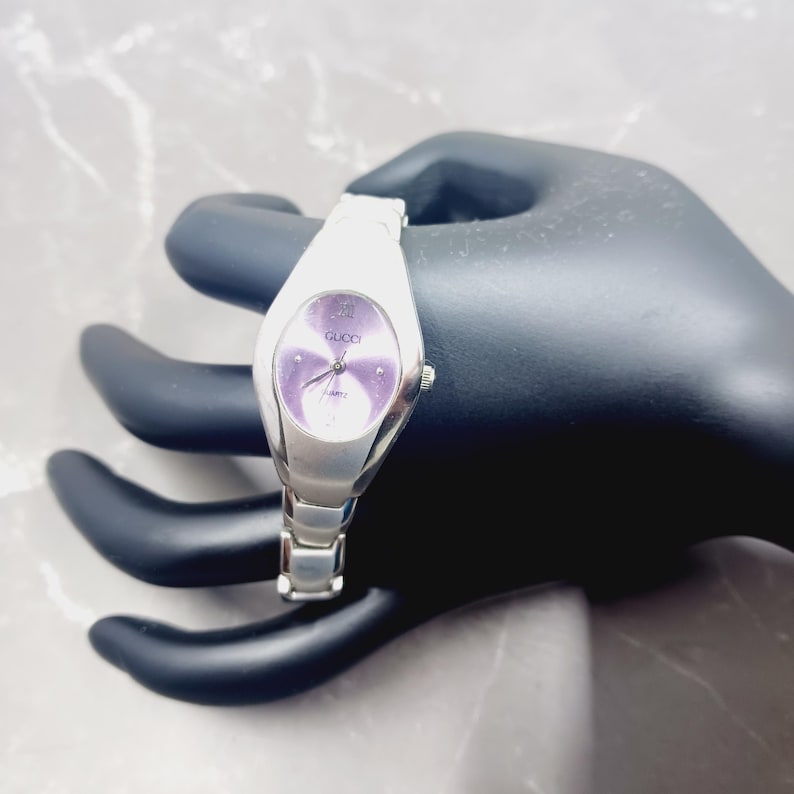 May include: A silver Gucci watch with a purple oval face. The watch has a silver strap and a quartz movement. The Gucci logo is visible on the watch face. The watch is displayed on a black surface.