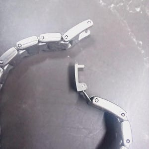 May include: Close-up of a silver watch strap with an open clasp. The strap is composed of connected rectangular links. The clasp mechanism is visible. The background is a dark, textured surface.