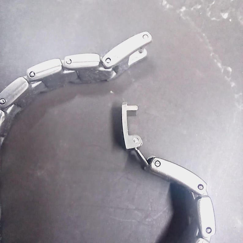 May include: Close-up of a silver watch strap with an open clasp. The strap is composed of connected rectangular links. The clasp mechanism is visible. The background is a dark, textured surface.