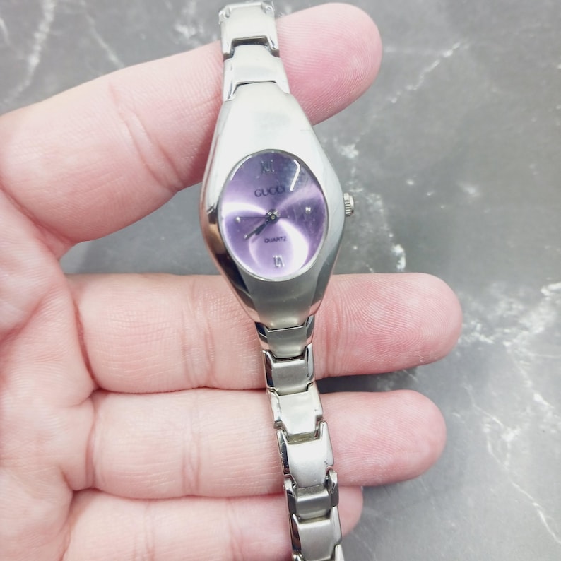 May include: A silver Gucci quartz watch with a purple oval face. The watch has silver hands and Roman numeral hour markers. The watch strap is made of silver links.